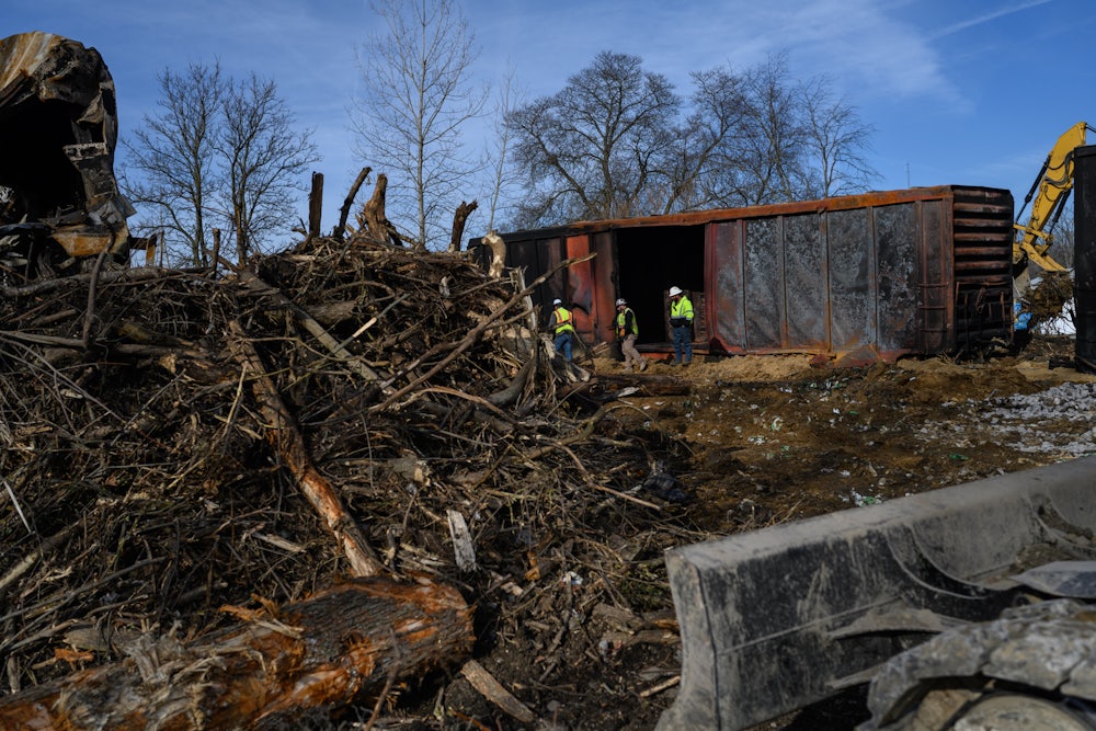 A cargo train, a giant pile of branches/uprooted trees, and three workers in the background.