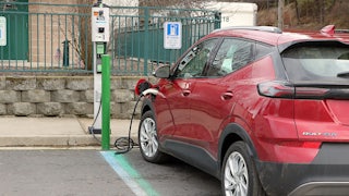 A red Chevrolet Volt is seen in a parking spot with a charging cord coming out of it to a charging station on the curb.