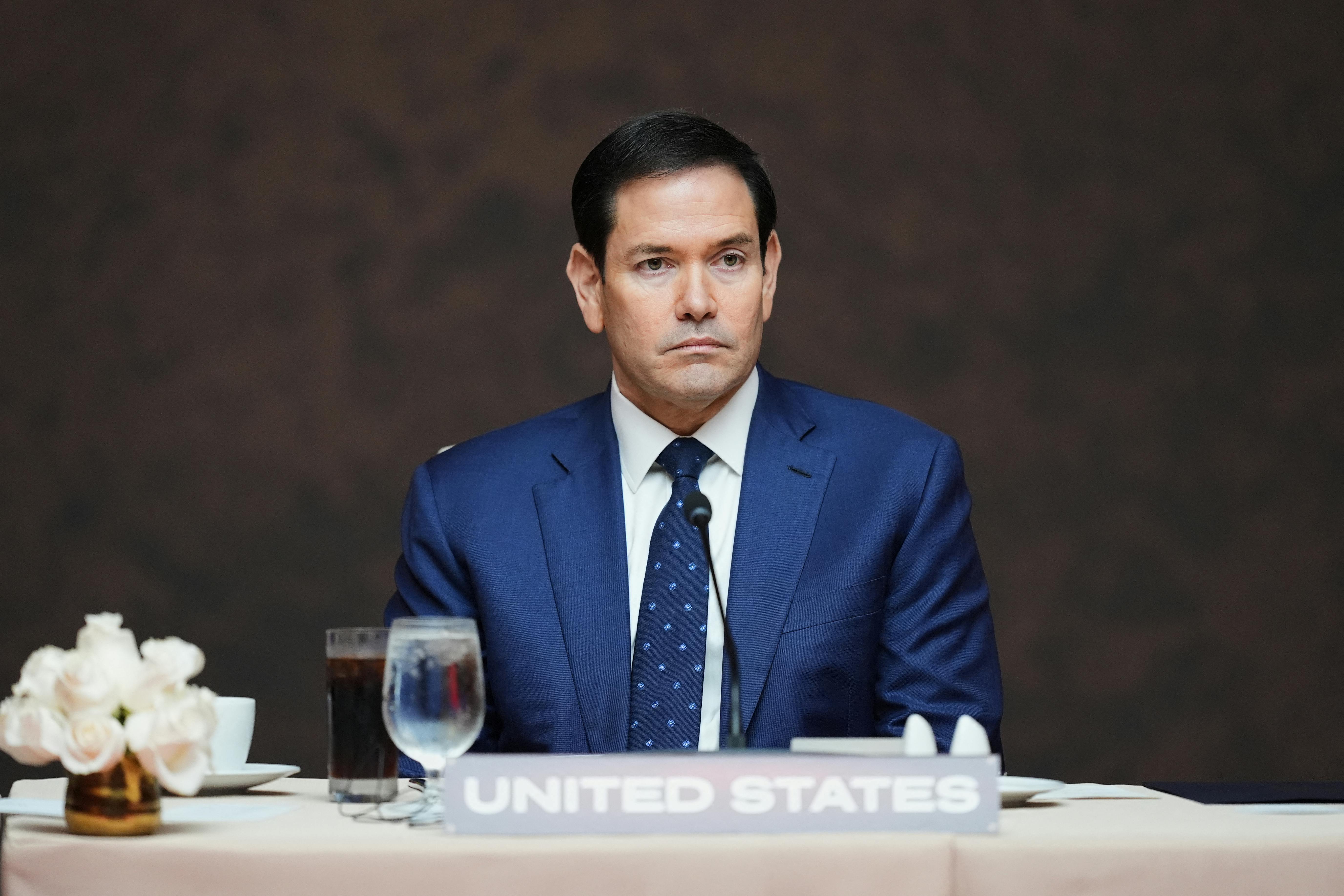 Marco Rubio looks uncomfortable as he sits at a table behind a United States nameplate.