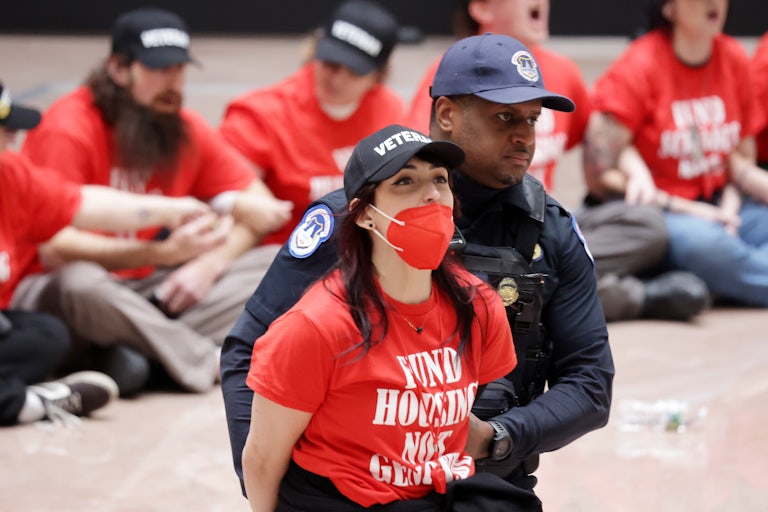A protester wearing a shirt reading "Fund Housing Not Genocide" is led out of the Capitol in handcuffs.