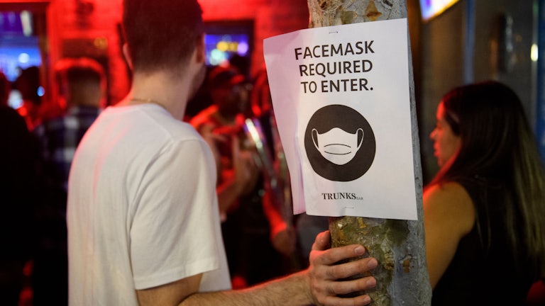 Unmasked people stand at the entrance of a bar, near a sign that reads: “Face masks required to enter.”