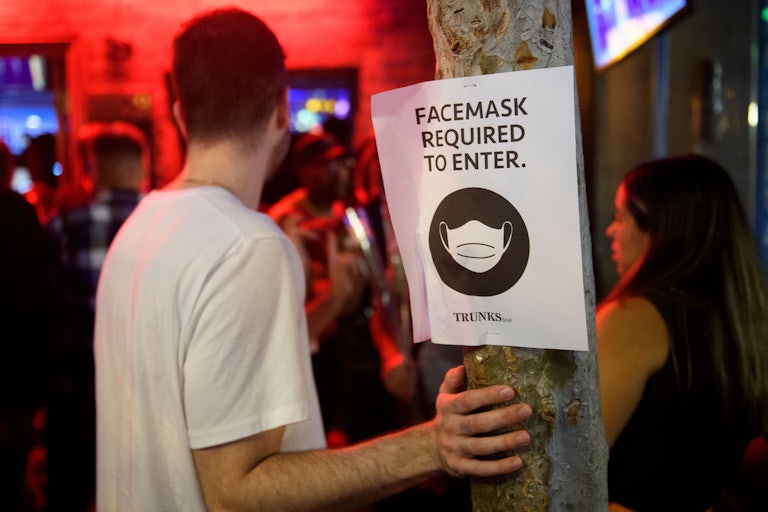 Unmasked people stand at the entrance of a bar, near a sign that reads: “Face masks required to enter.”