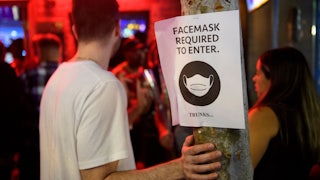 Unmasked people stand at the entrance of a bar, near a sign that reads: “Face masks required to enter.”