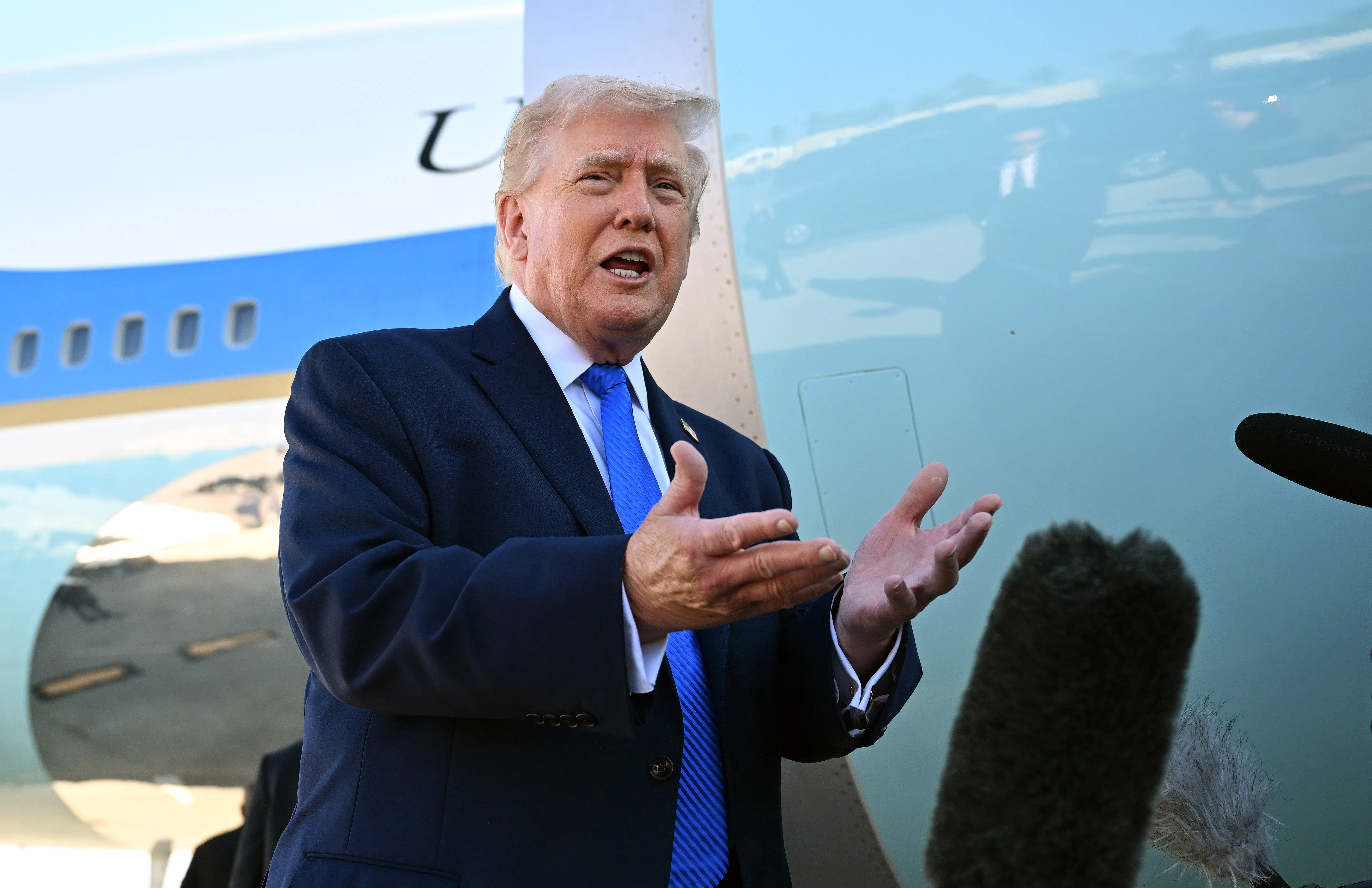 Donald Trump, wearing a blue suit and blue tie, stands with his hands open in front of Air Force One.