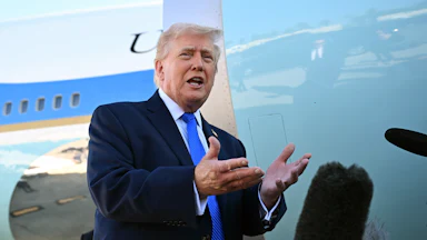 Donald Trump, wearing a blue suit and blue tie, stands with his hands open in front of Air Force One.