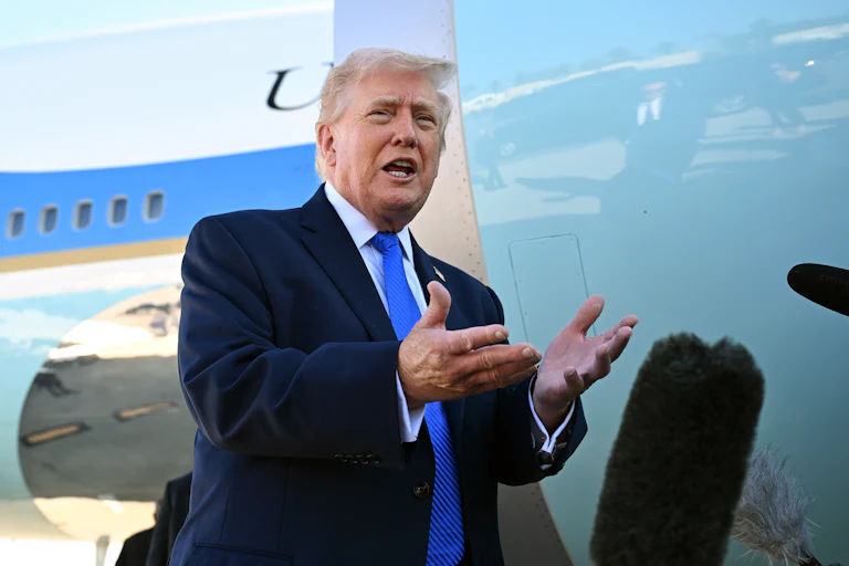Donald Trump, wearing a blue suit and blue tie, stands with his hands open in front of Air Force One.