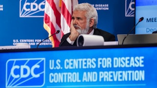 Robert Malone rests his chin in his hand while surrounded by blue "CDC" banners.