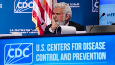 Robert Malone rests his chin in his hand while surrounded by blue "CDC" banners.