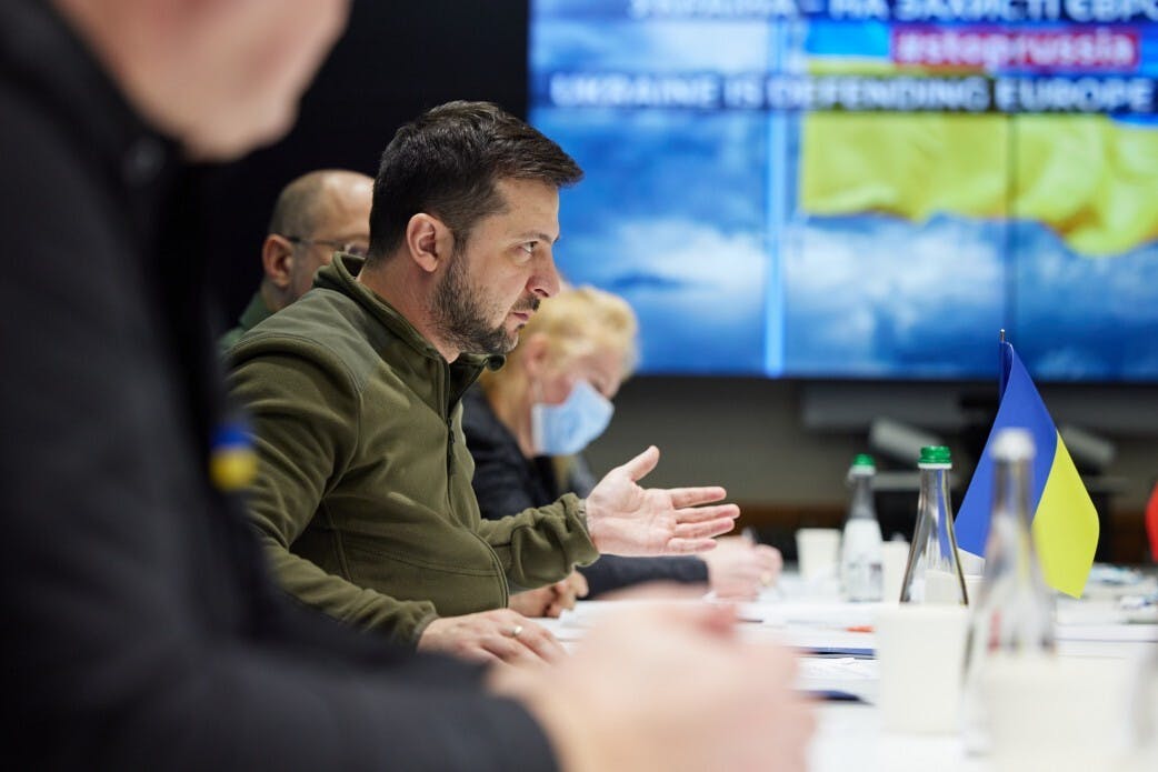 Volodymyr Zelenskiy sits at a table with others, gesturing while speaking.