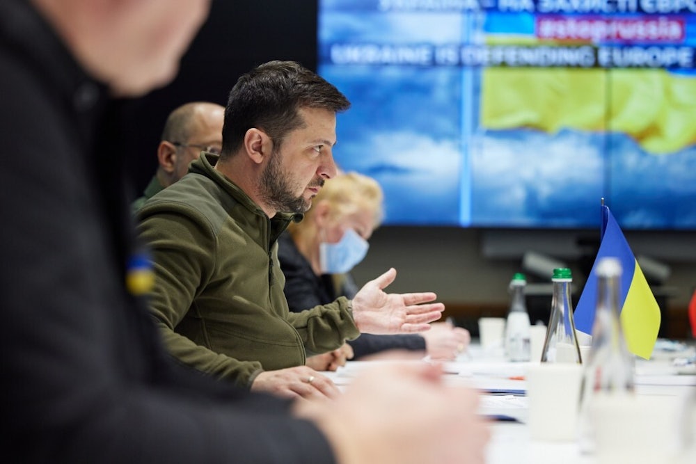 Volodymyr Zelenskiy sits at a table with others, gesturing while speaking.