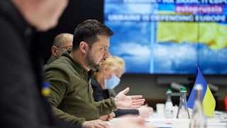 Volodymyr Zelenskiy sits at a table with others, gesturing while speaking.
