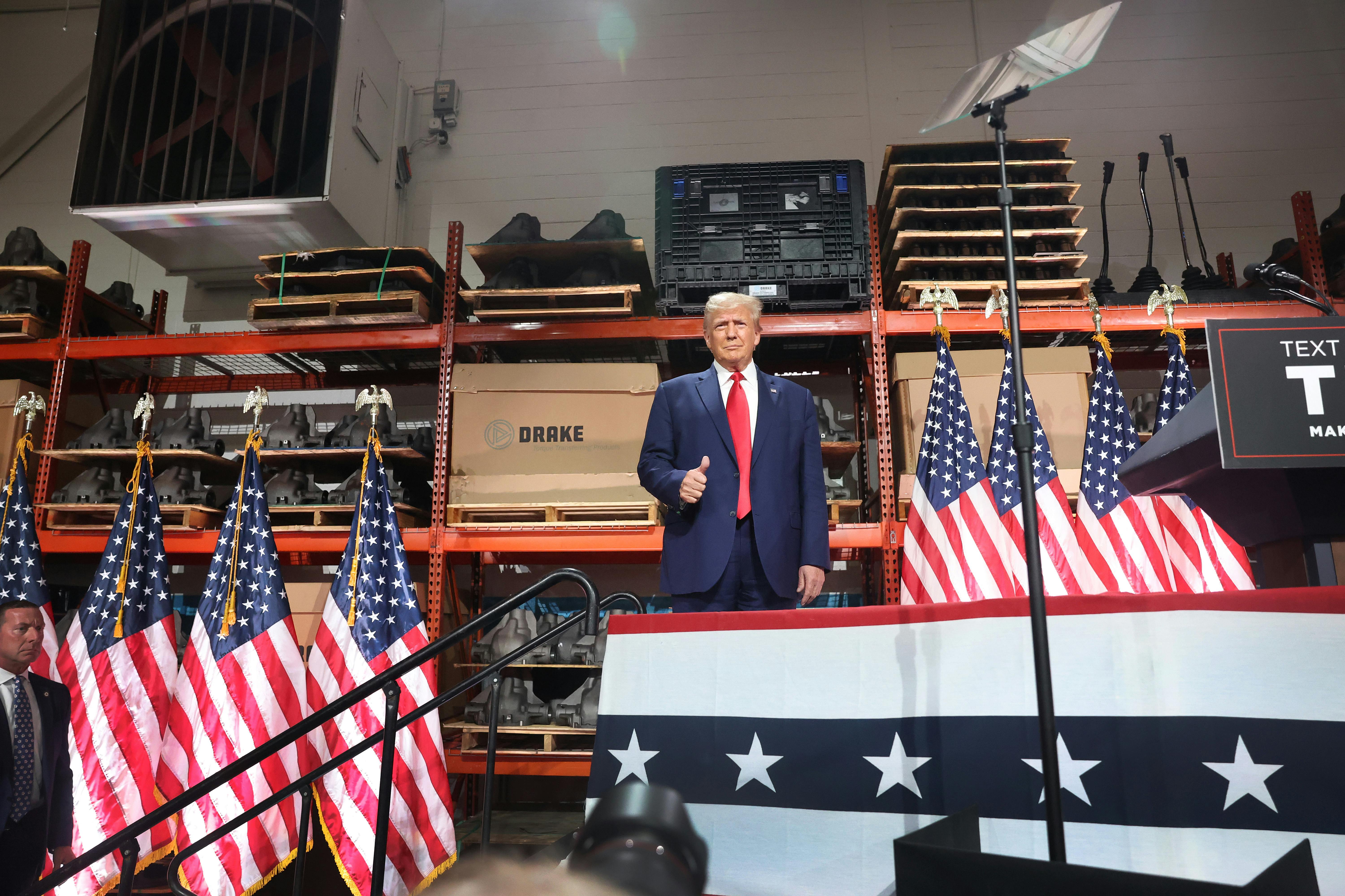 Trump stands on a platform bedecked with American flags in front of automotive parts.