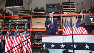 Trump stands on a platform bedecked with American flags in front of automotive parts.