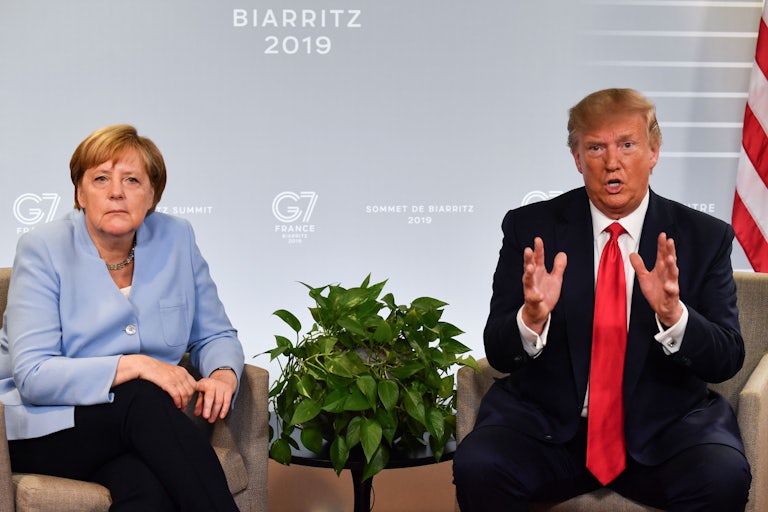 Angela Merkel sits next to Donald Trump while he gestures and speaks