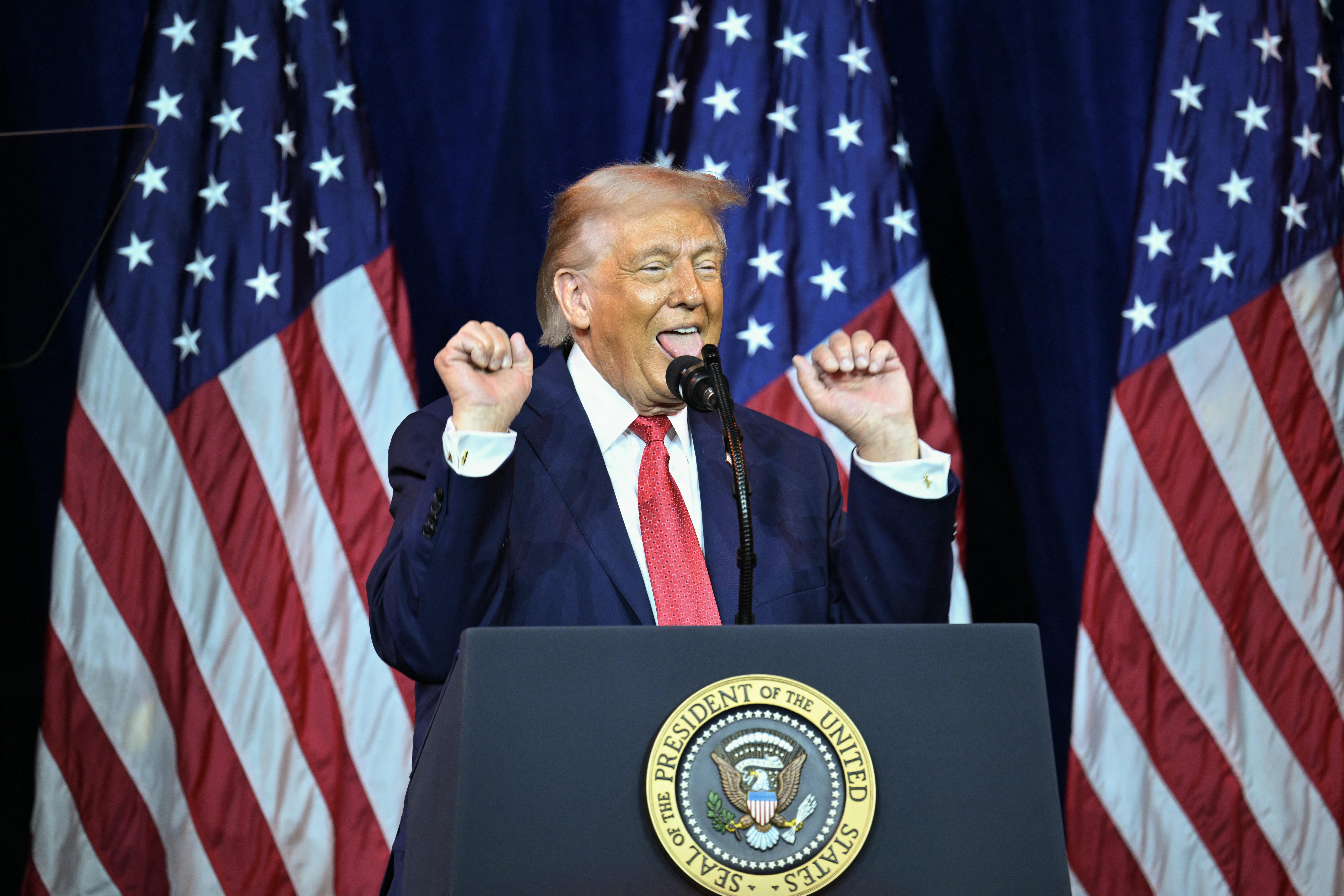 Donald Trump raises his fists and sticks out his tongue while standing at a podium