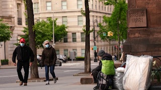 An unhoused person sitting on the sidewalk in Manhattan as pedestrians pass.