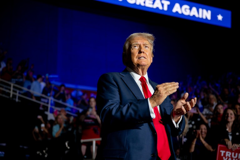 Donald Trump smiles weirdly and claps. A crowd is seen behind him.