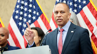 Hakeem Jeffries gesticulates while speaking at a lectern, with other people and American flags behind him.