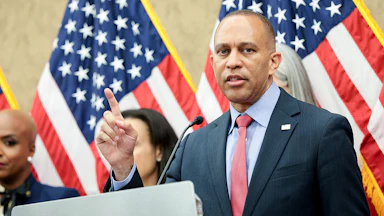 Hakeem Jeffries gesticulates while speaking at a lectern, with other people and American flags behind him.