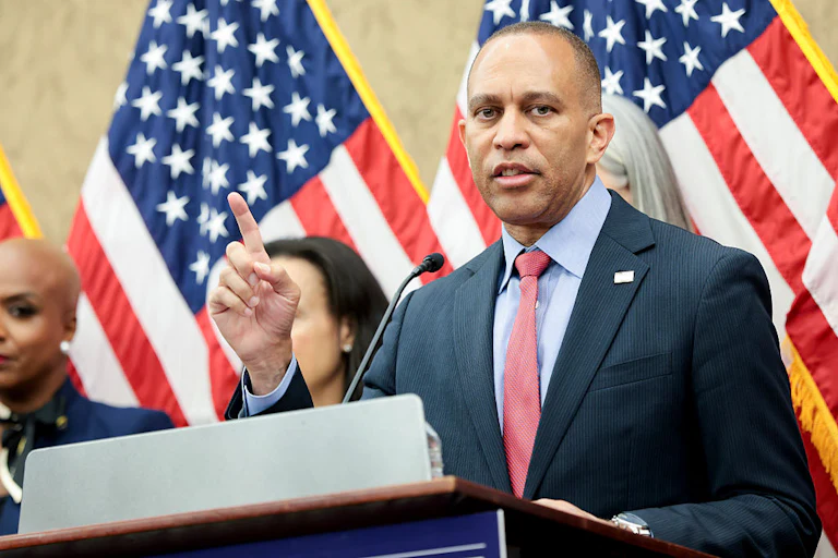 Hakeem Jeffries gesticulates while speaking at a lectern, with other people and American flags behind him.