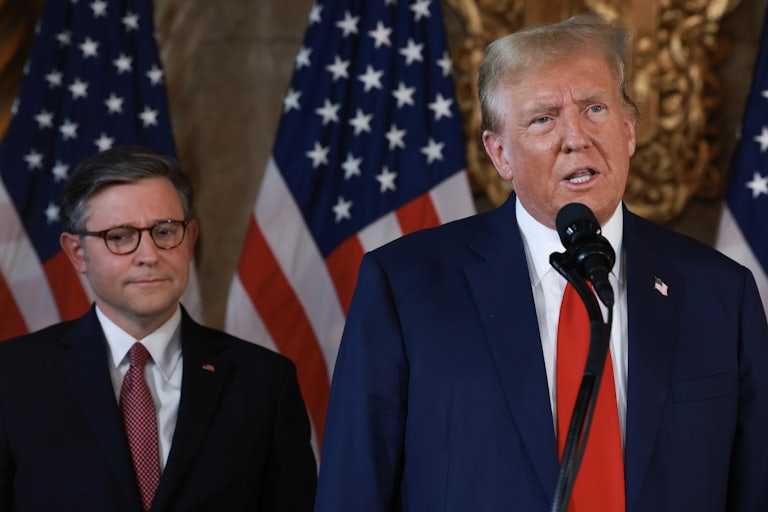 Donald Trump speaks into a microphone while Mike Johnson stands behind him and frowns