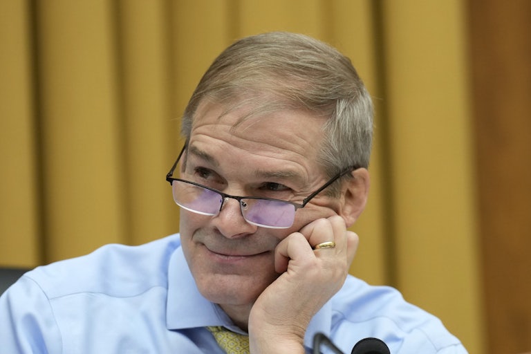 Jim Jordan, wearing glasses, does a coy little grin as he holds his head in his hands in front of a gold backdrop