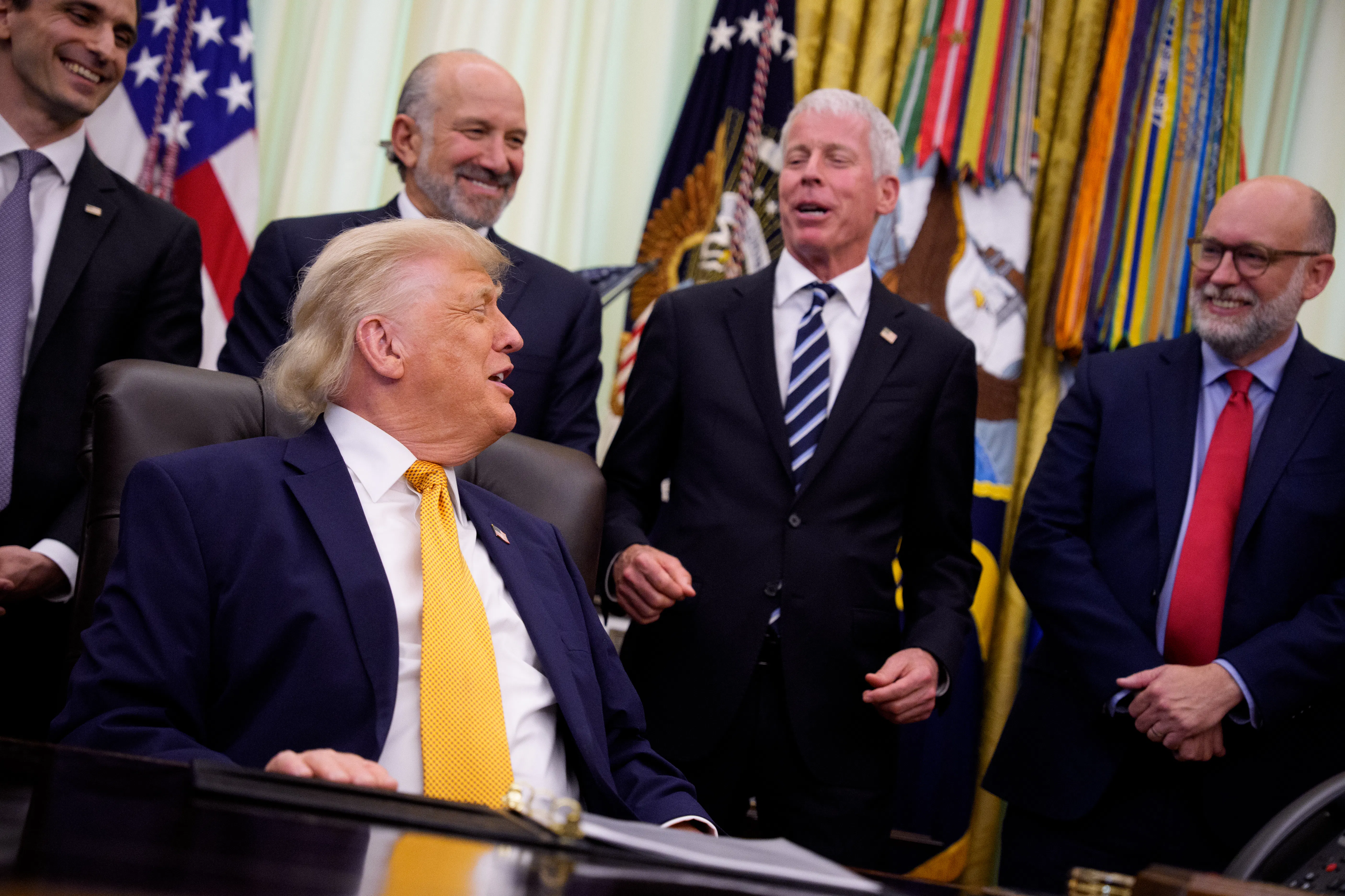 President Donald Trump laughs while seated at his desk in the Oval Office of the White House, while Office of Management and Budget Director Russell Vought, Energy Secretary Chris Wright, Commerce Secretary Howard Lutnick, and White House Office of Science and Technology Policy Director Michael Kratsios stand behind him.