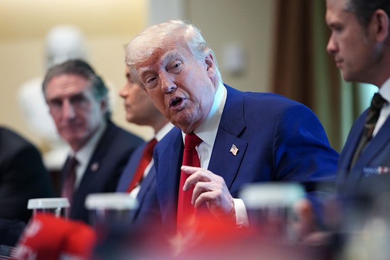 Donald Trump sits at a conference table speaking to his Cabinet. In front of each person is a red MAGA cap.