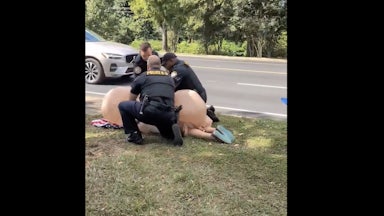 Three police officers (two white, one Black) arrest a woman dressed in an inflatable penis costume, as she lays on her face on the ground.