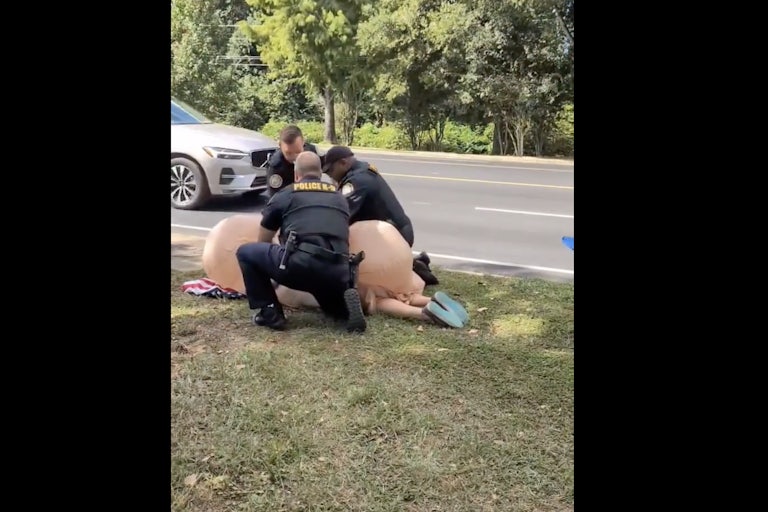 Three police officers (two white, one Black) arrest a woman dressed in an inflatable penis costume, as she lays on her face on the ground.