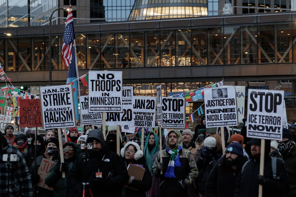 A sea of protesters standing in dim outdoor light hold signs with messages like "No Troops in Minnesota" and "Stop Ice Terror Now!"