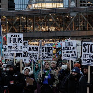 A sea of protesters standing in dim outdoor light hold signs with messages like "No Troops in Minnesota" and "Stop Ice Terror Now!"