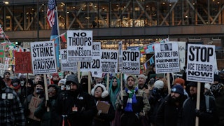 A sea of protesters standing in dim outdoor light hold signs with messages like "No Troops in Minnesota" and "Stop Ice Terror Now!"