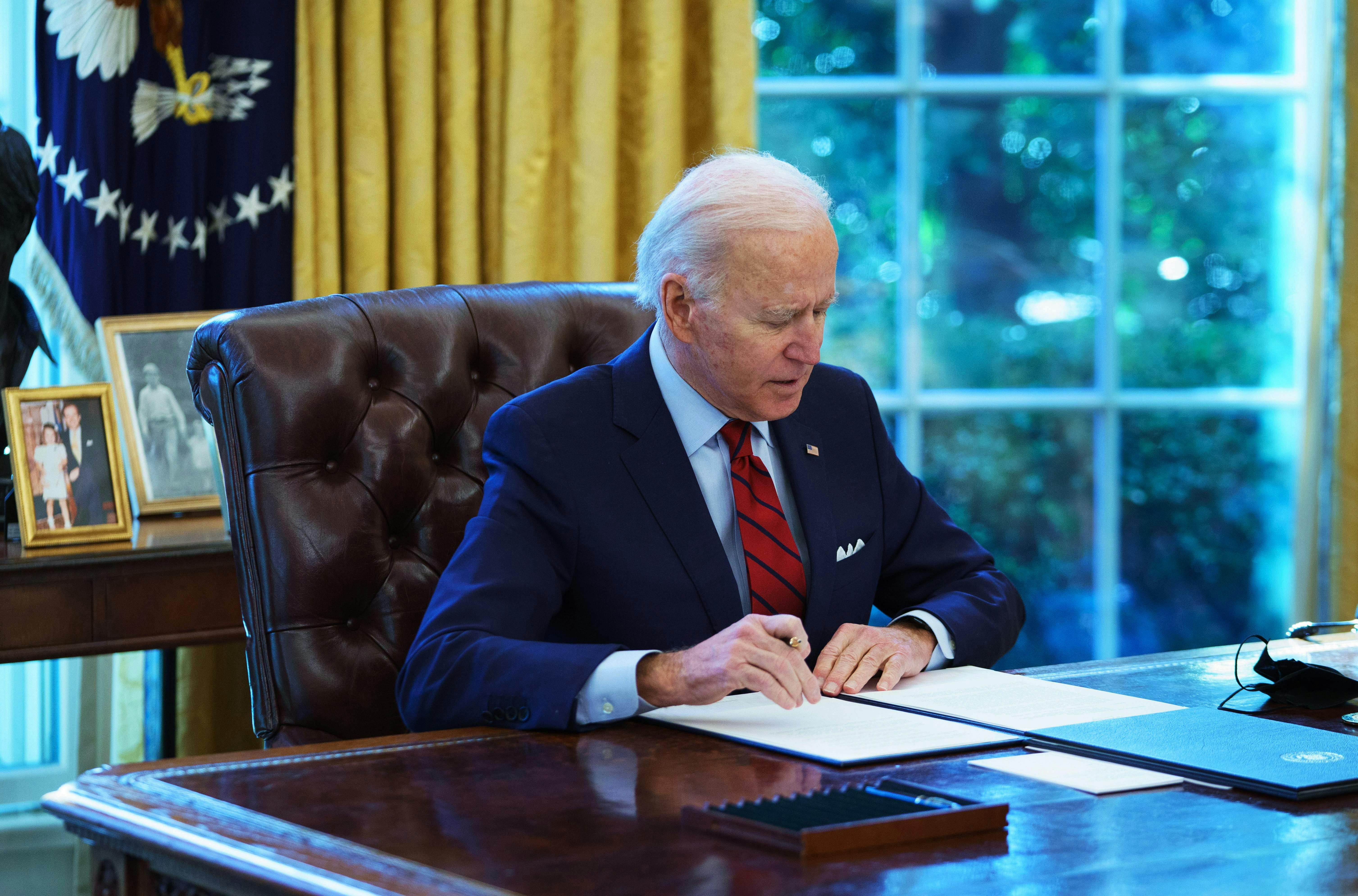 A seated Joe Biden holds a pen in his right hand while preparing to sign an executive order in the Oval Office 