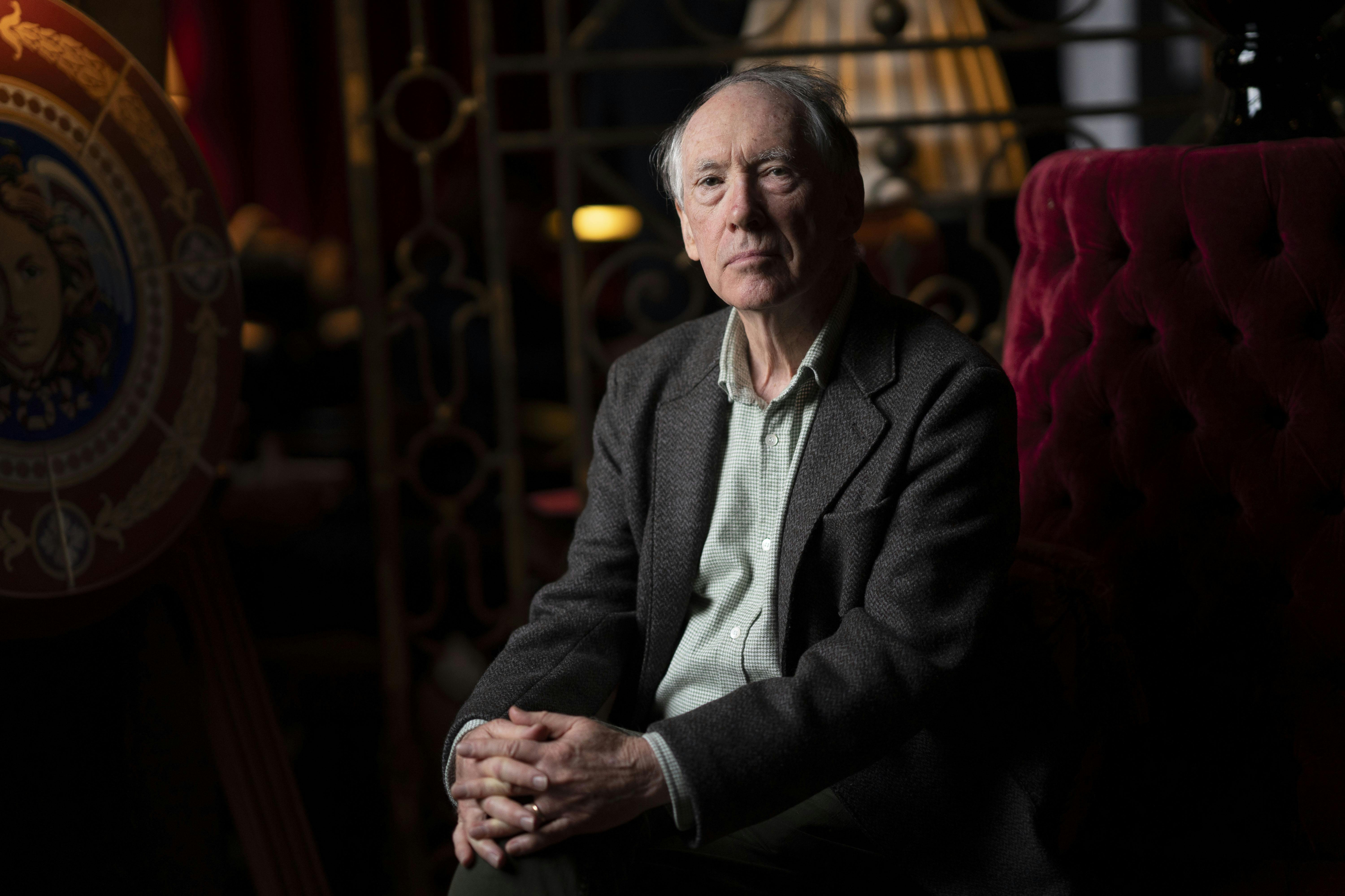 Ian McEwan sitting in a dark room on a red chair with his handed folded together.