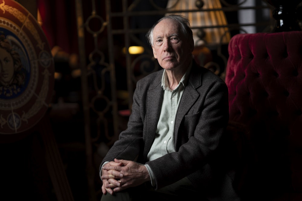 Ian McEwan sitting in a dark room on a red chair with his handed folded together.