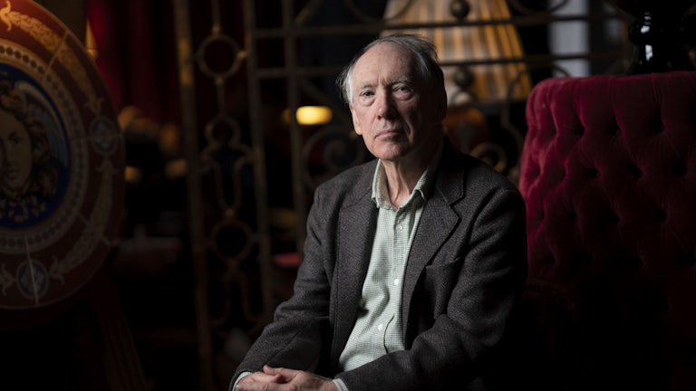 Ian McEwan sitting in a dark room on a red chair with his handed folded together.