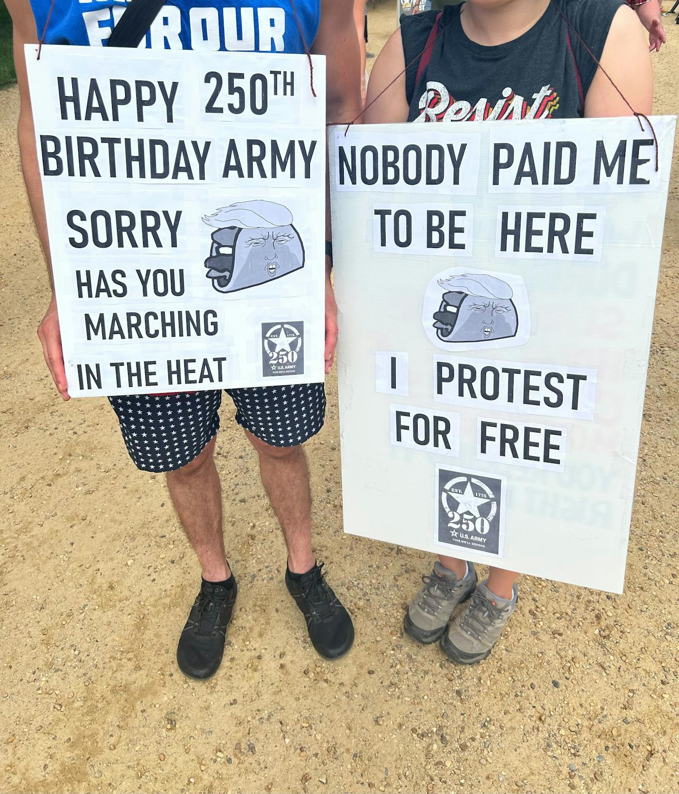 Two people hold signs reading "Happy 250th Birthday Army, Sorry [image of taco] Has You Marching In The Heat" and "Nobody Paid Me to Be Here I Protest for Free." Both signs have taco images that are made to look like Donald Trump.