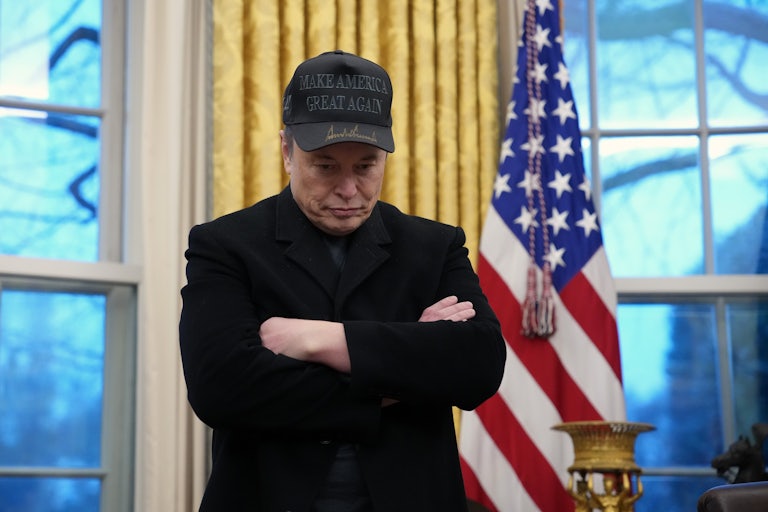 Elon Musk crosses his arms and looks downward while standing in the Oval Office.