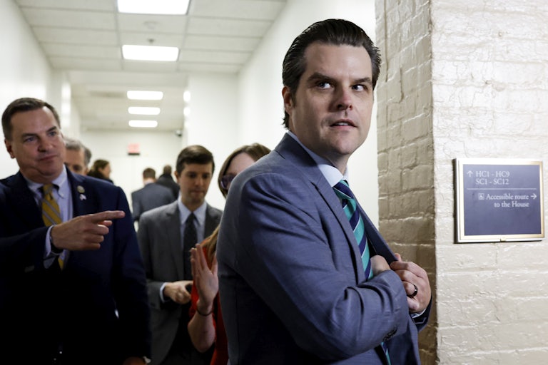 Matt Gaetz puts a hand in his suit pocket and looks off camera. Others surround him in the background; one points at him.