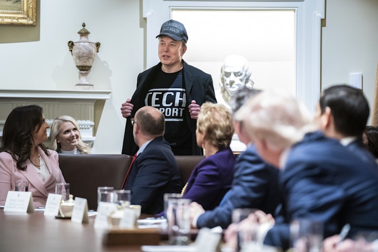 Elon Musk holds open his jacket to reveal the words "Tech Support" on his T-shirt, as Donald Trump and other Cabinet members look at him