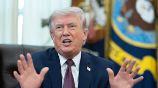 Donald Trump holds both hands up and speaks while sitting at his desk in the Oval Office