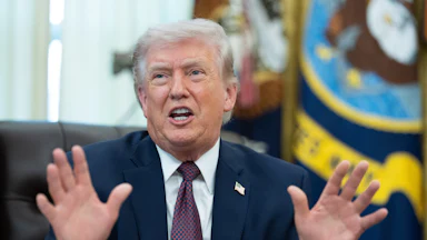 Donald Trump holds both hands up and speaks while sitting at his desk in the Oval Office