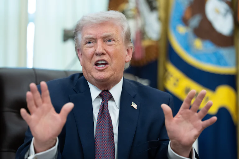 Donald Trump holds both hands up and speaks while sitting at his desk in the Oval Office