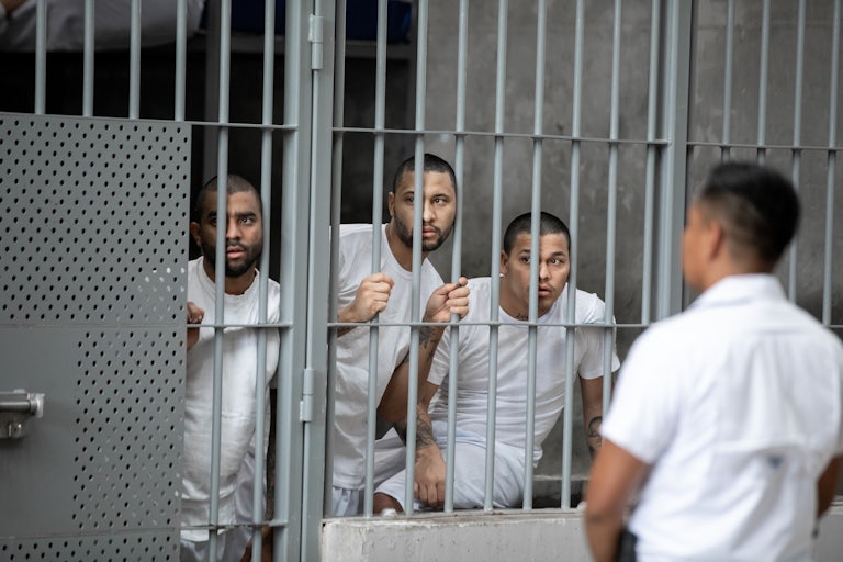 Three men deported from the United States imprisoned in CECOT in El Salvador look out from behind the bars in their cell.