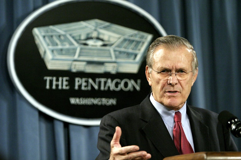 Donald Rumsfeld speaks at a podium with his right hand raised in front of a blue Pentagon backdrop in 2005.