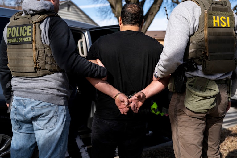 Two federal agents wearing flak jackets hold the arms of a detained immigration suspect wearing a black t-shirt with his hands handcuffed behind his back.
