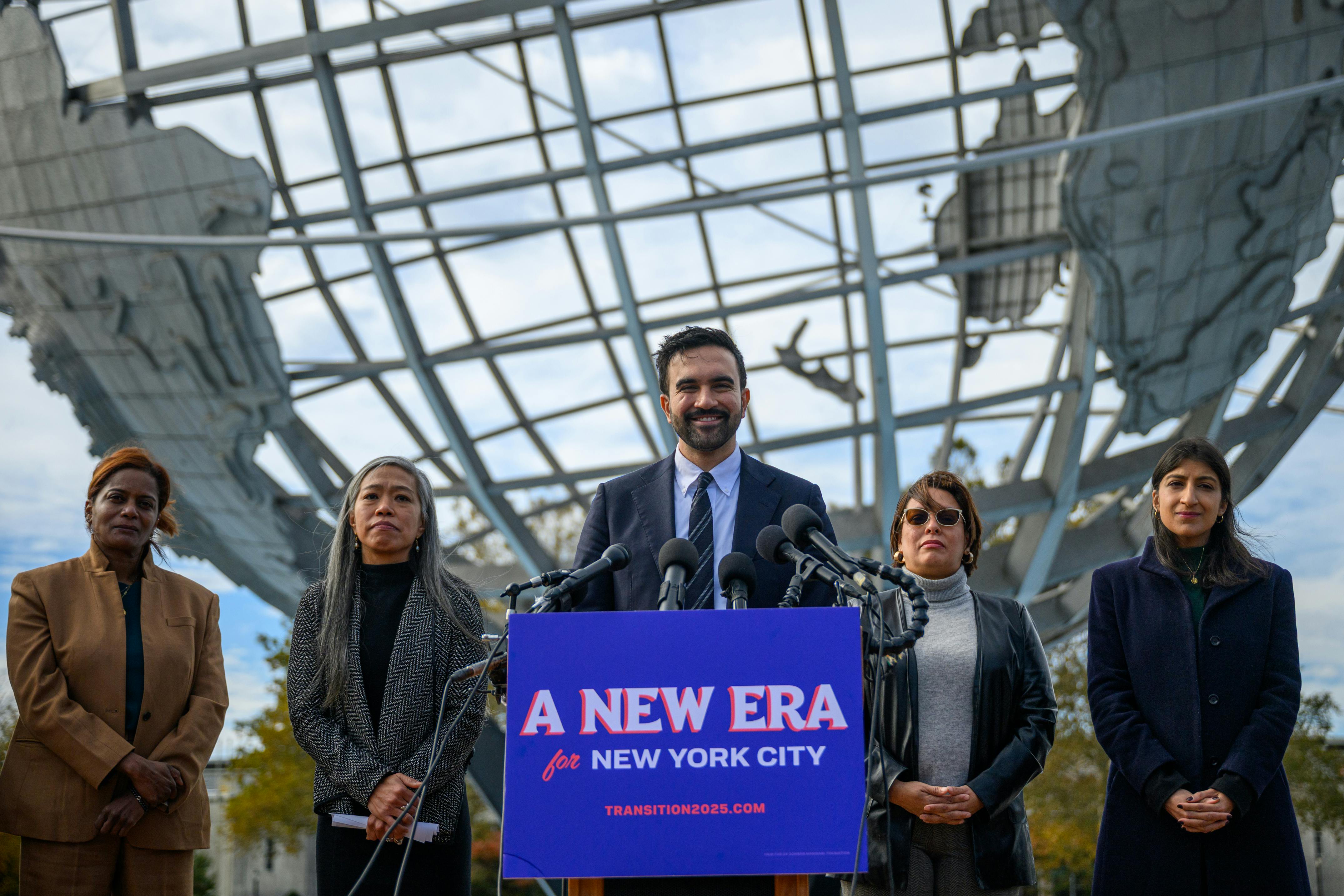 New York City Mayor-elect Zohran Mamdani smiles while standing at a podium with a sign that says "A new era for New York City." He is flanked by his transition team members Melanie Hartzog, Maria Torres-Springer, Grace Bonilla and Lina Khan