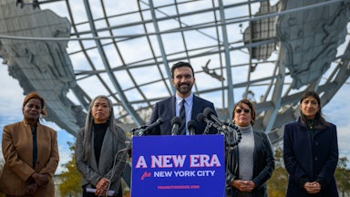 New York City Mayor-elect Zohran Mamdani smiles while standing at a podium with a sign that says "A new era for New York City." He is flanked by his transition team members Melanie Hartzog, Maria Torres-Springer, Grace Bonilla and Lina Khan