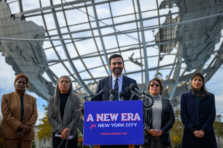New York City Mayor-elect Zohran Mamdani smiles while standing at a podium with a sign that says "A new era for New York City." He is flanked by his transition team members Melanie Hartzog, Maria Torres-Springer, Grace Bonilla and Lina Khan